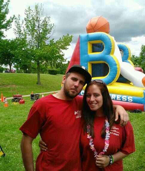 A man and a woman in matching red shirts stand in a park in front of an inflatable basketball game.