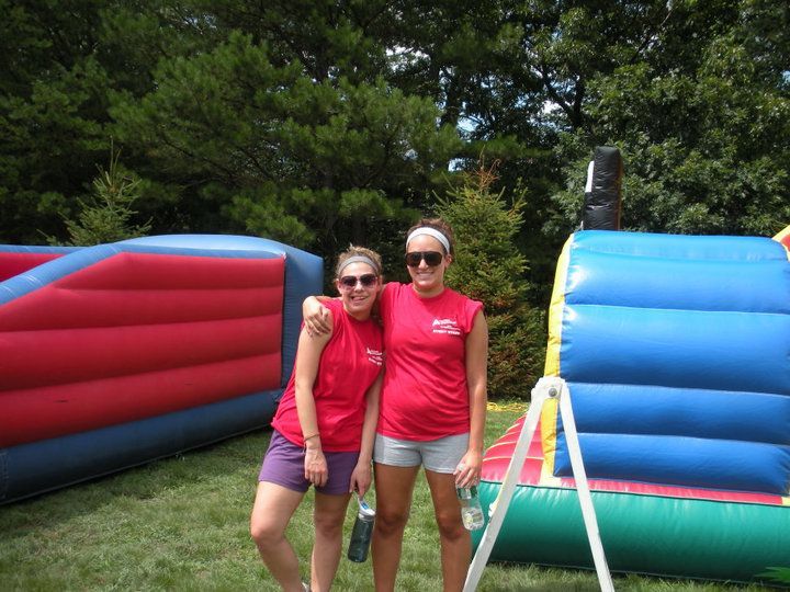 Two people in matching red shirts and shorts stand smiling between two large, colorful inflatable structures on grass.