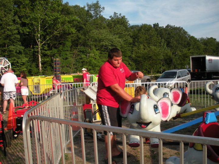 A person in a red shirt stands next to an elephant-shaped carnival ride at an outdoor fair.