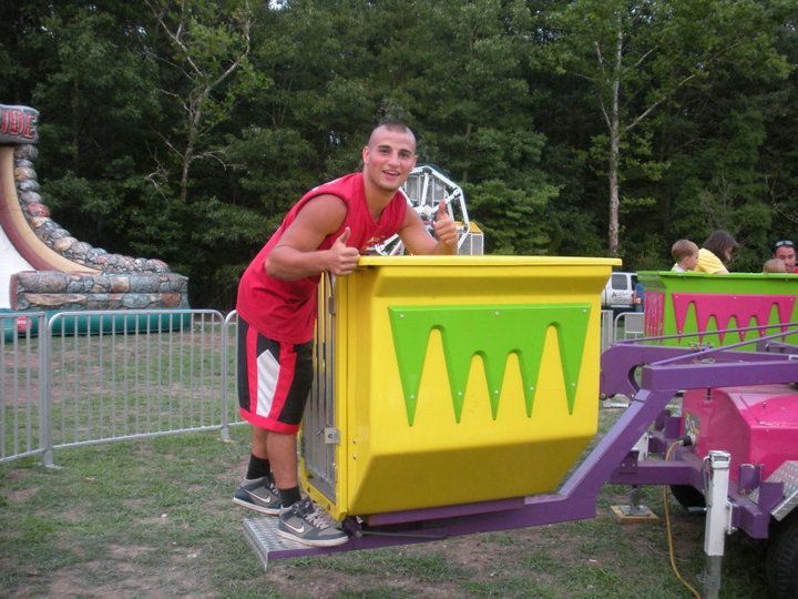 A person in a red tank top and shorts stands next to a bright yellow ride car at a carnival, giving a thumbs-up.