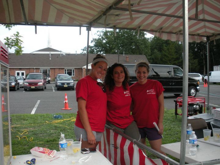 Three people in red shirts stand smiling under a striped canopy at an outdoor event near a parking lot.