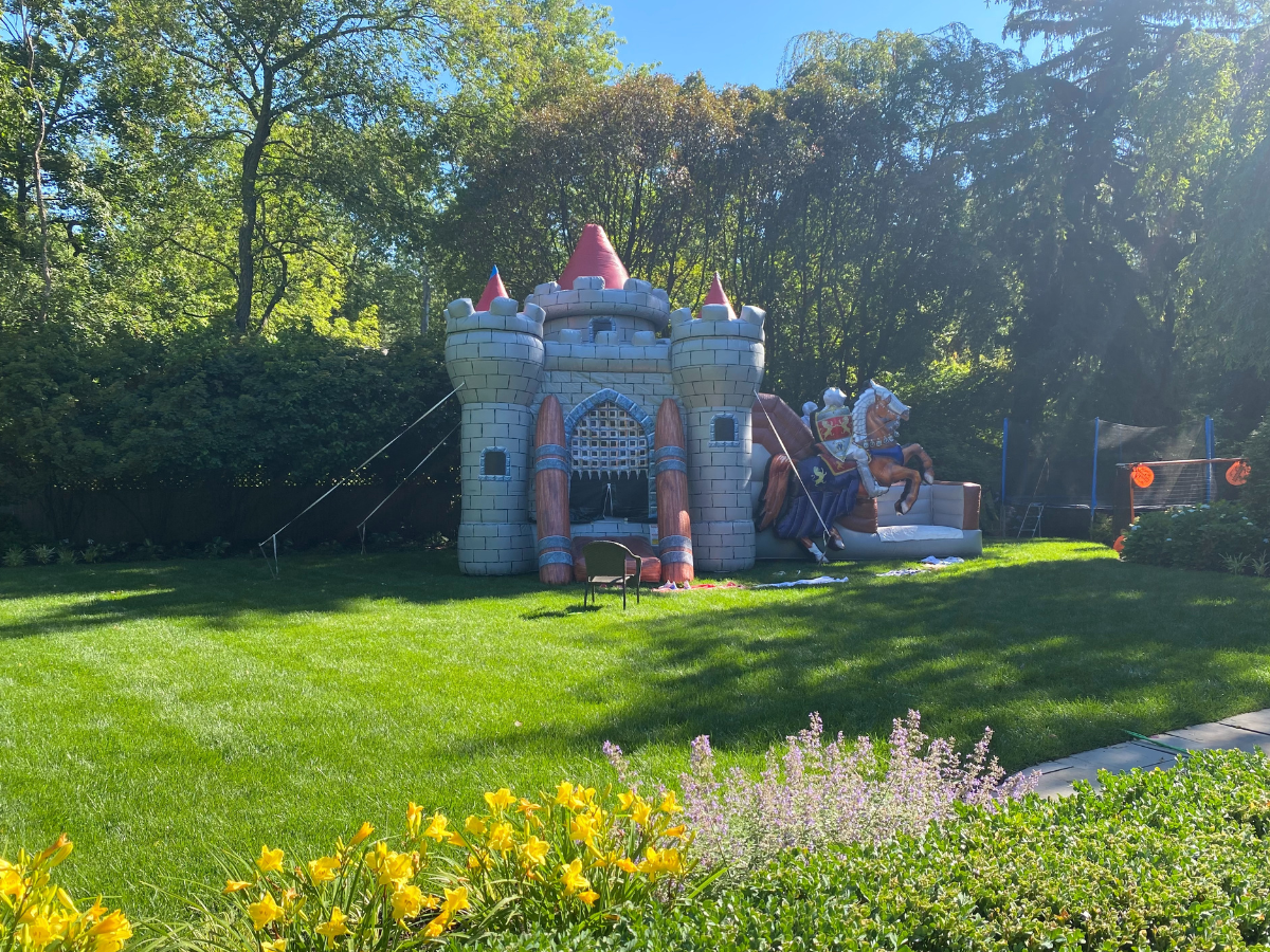 An inflatable grey castle bounce house sits in a sunny backyard with green grass and trees in the background.