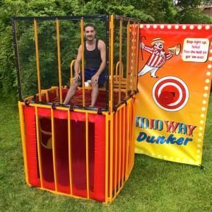 A person sitting in a yellow-and-red portable dunk tank next to a banner with a target and the text 
