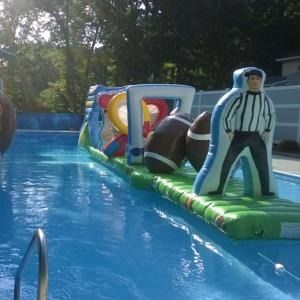 An inflatable football-themed obstacle course floats in a swimming pool, featuring a referee figure and footballs.