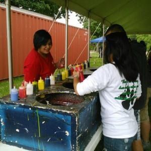 Two people stand at a craft station under a tent, preparing to dye fabric in tubs of colorful liquid.