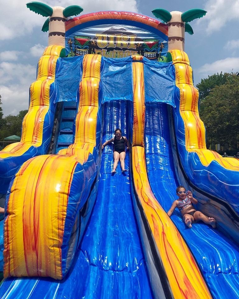 Two people slide down a tall, blue and yellow inflatable water slide on a sunny day.