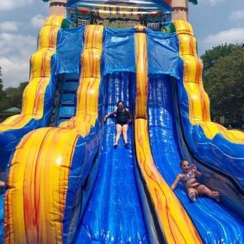 Two people slide down a tall, blue and yellow inflatable water slide on a sunny day.