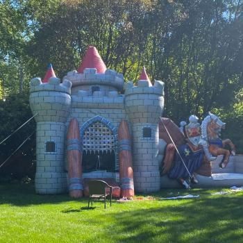 An inflatable castle bounce house with a knight and horse theme, set up on a green lawn in a wooded area.