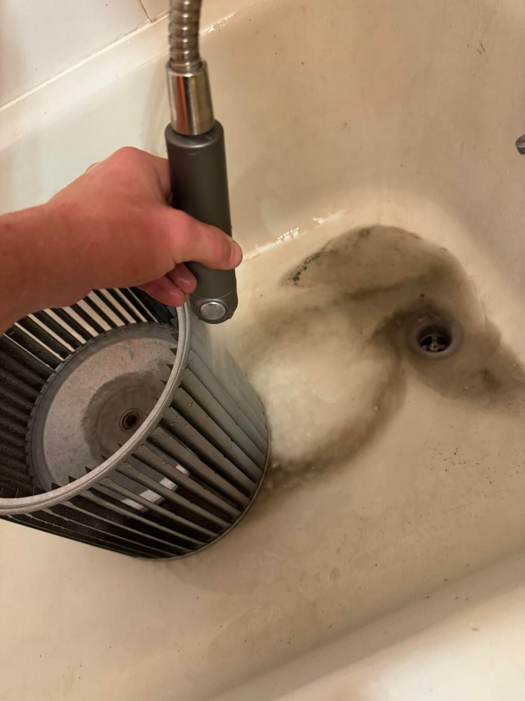 A person cleaning a dirty, grey circular HVAC blower wheel with a handheld shower sprayer in a bathtub.