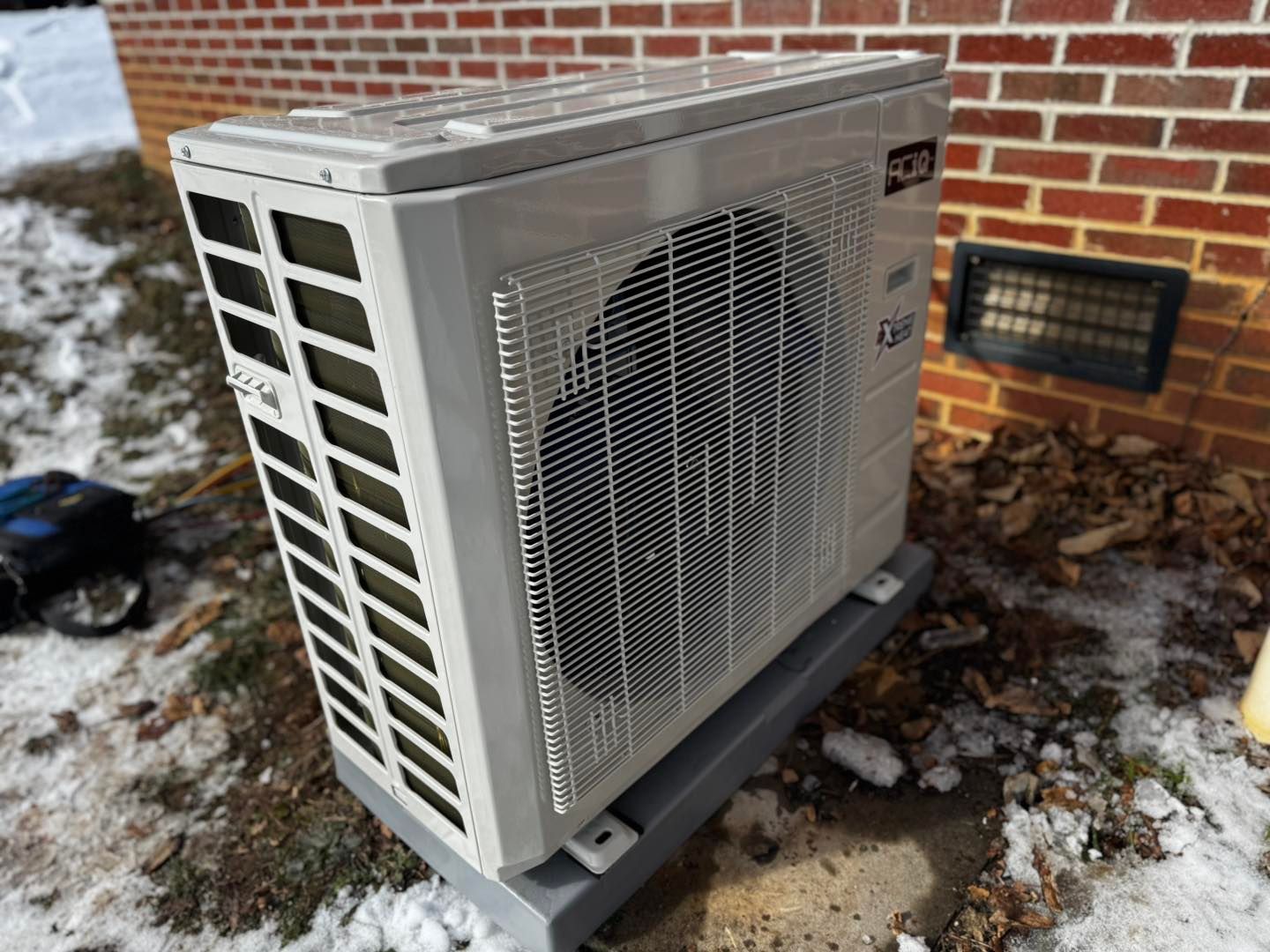 An outdoor HVAC heat pump unit sits on a concrete pad against a brick wall, with patches of snow on the ground nearby.