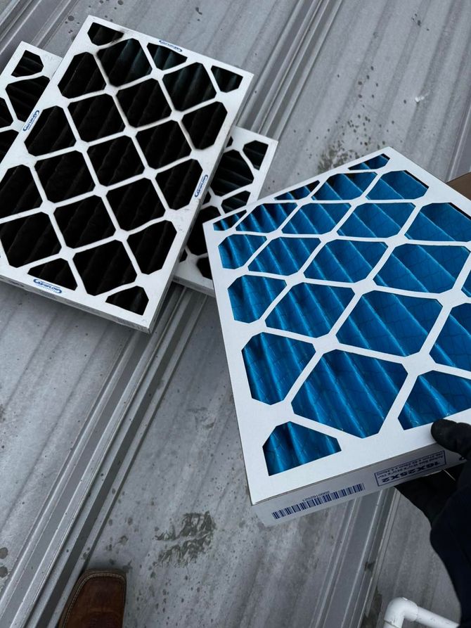 A hand in a black glove holds a blue HVAC filter next to used, black filters on a metal roof.