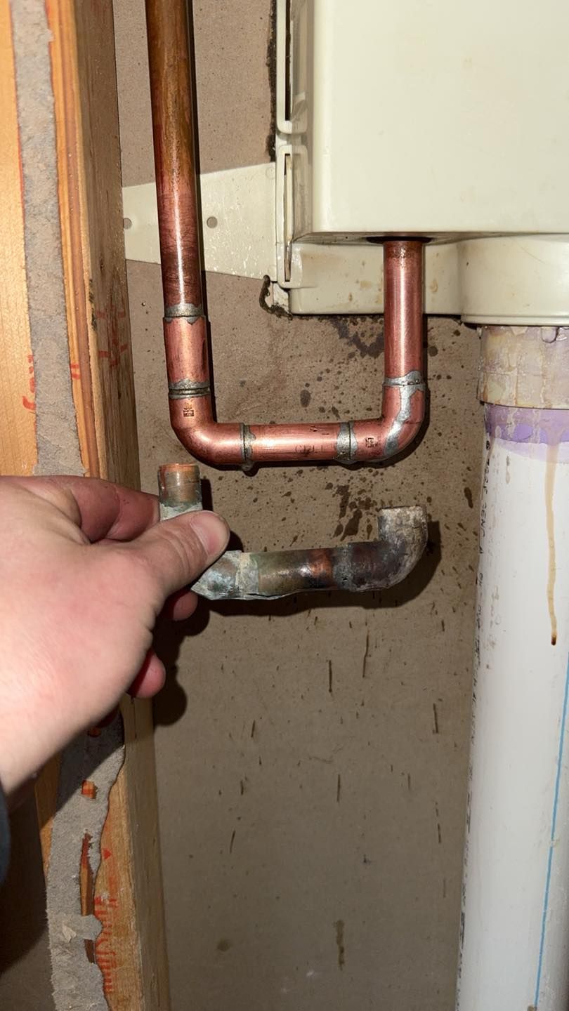 A hand holds a severed section of copper piping beneath an installed U-shaped pipe fitting attached to a wall heater.