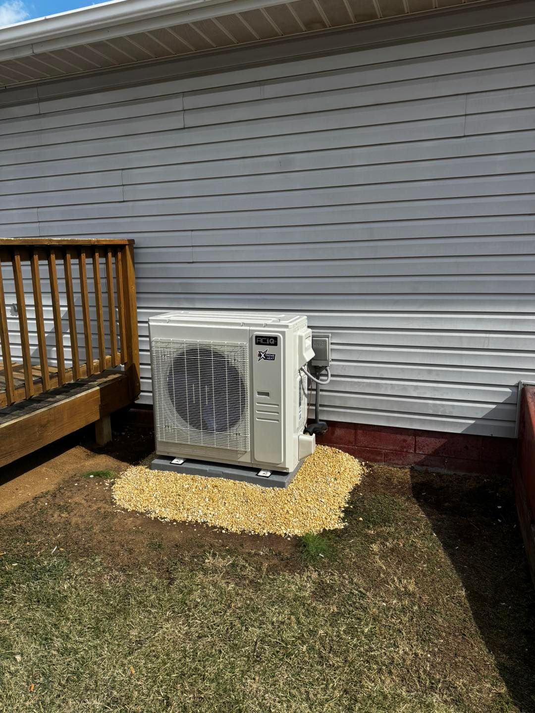 A beige HVAC outdoor unit sits on a small gravel pad next to a light-colored vinyl-sided house and a wooden deck.