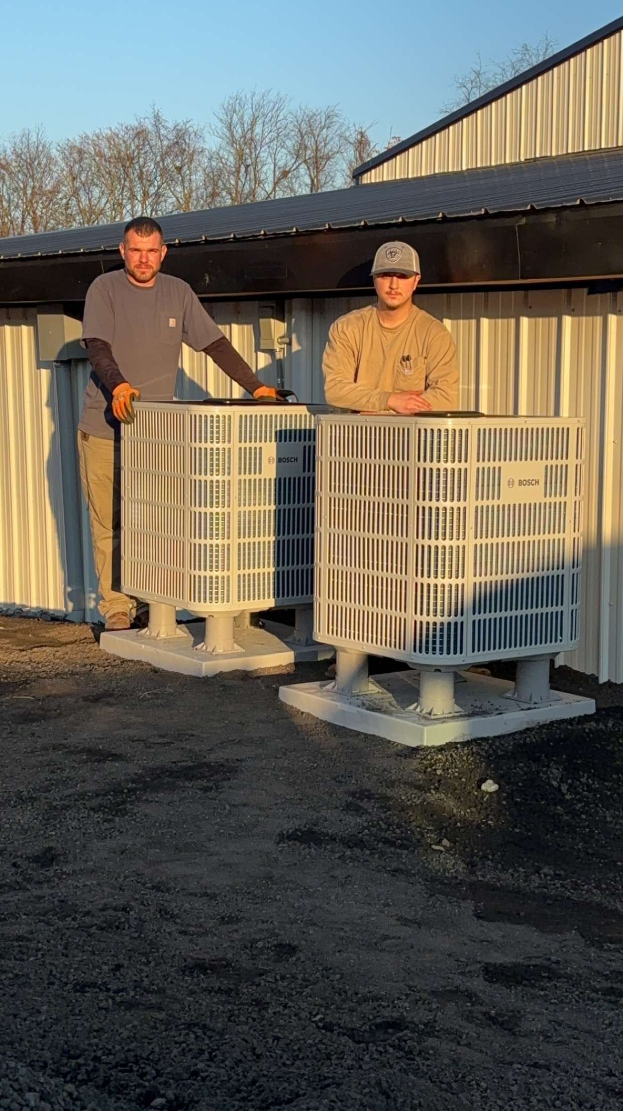 Two people stand outdoors behind two large, white, square industrial containers on concrete bases in front of a shed.