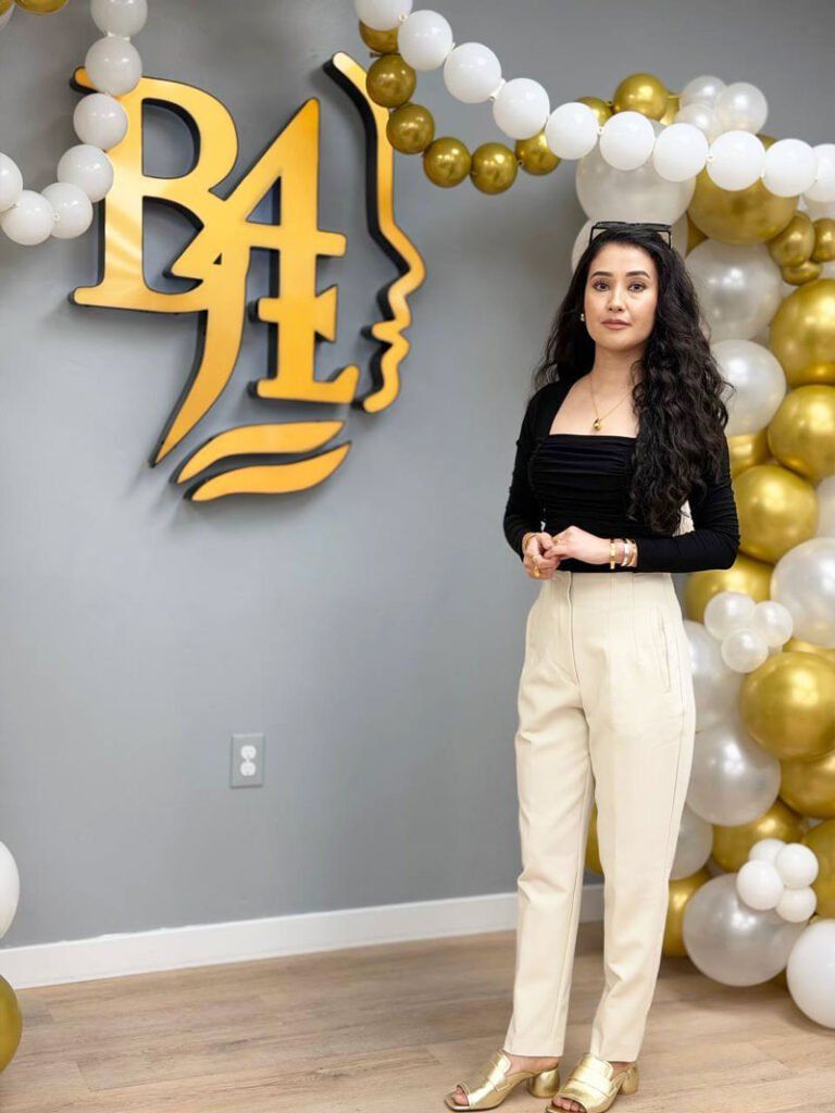 Woman standing beside a gold-and-white balloon arch and gold wall letters in a decorated room.