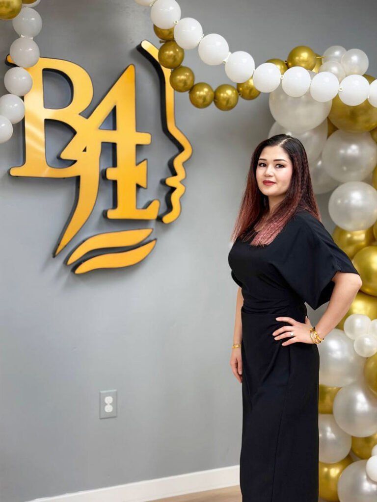 Woman in black dress posing beside gold-and-white balloon backdrop and gold wall logo