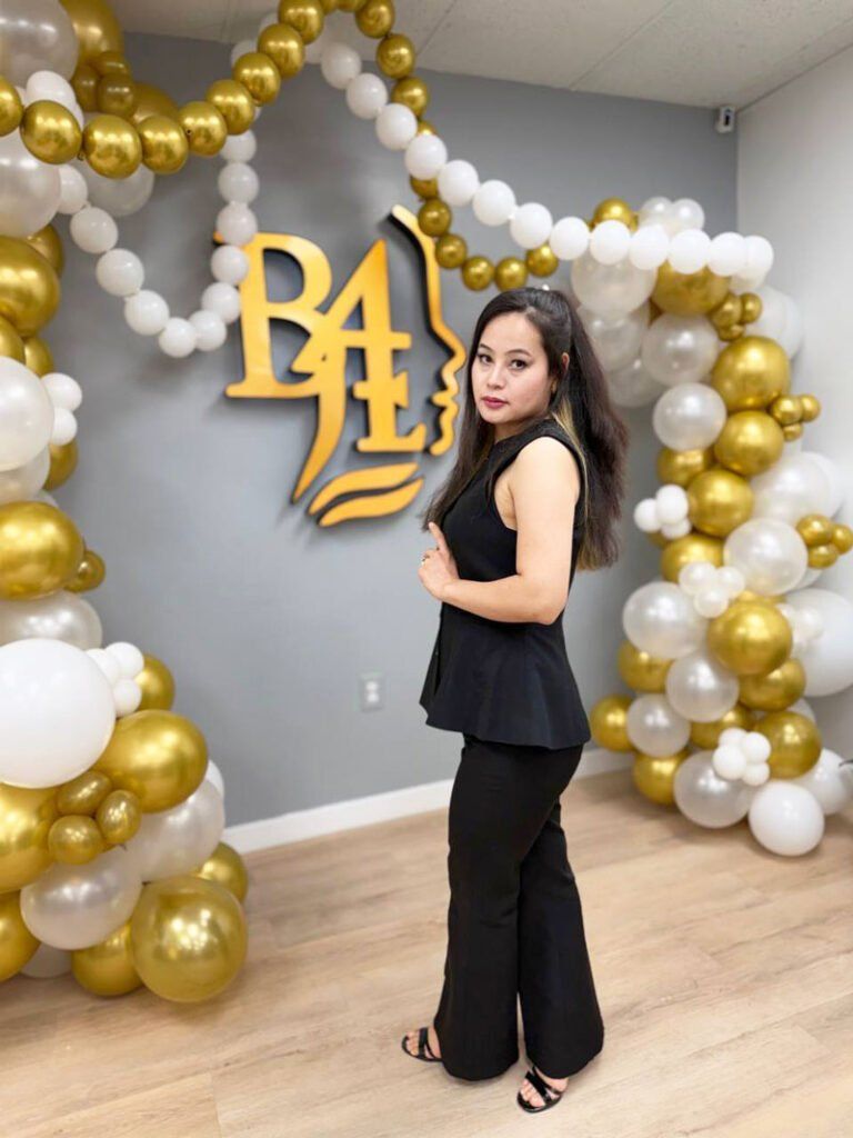 Woman in black posing beside gold-and-white balloon arch and gold monogram backdrop at an indoor celebration