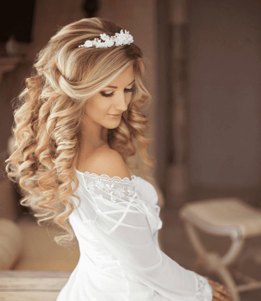 Bride in a white off-the-shoulder gown with curled blonde hair and floral headpiece, looking down indoors