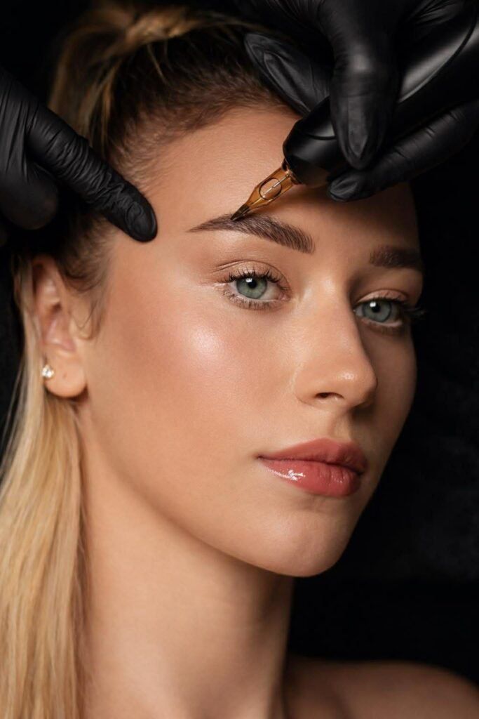 Close-up of a woman receiving eyebrow grooming with gloved hands against a dark background