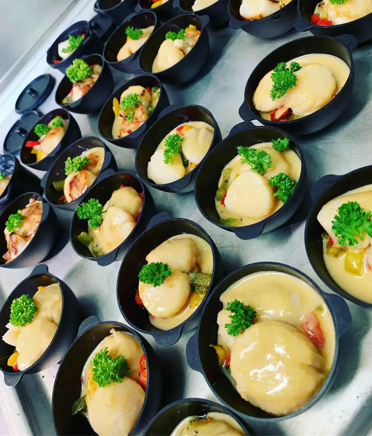 De petits bols noirs remplis de mets préparés, probablement des hors-d'œuvre, sont disposés en rangées. Les plats contiennent des raviolis, de la sauce et des légumes, le tout agrémenté de persil.