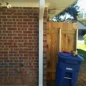 A blue trash can is sitting on the side of a brick wall next to a wooden fence.