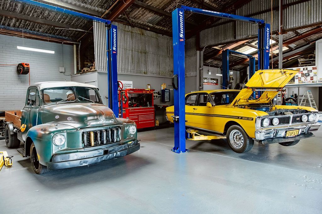 Two vintage cars in an auto shop, one green and one yellow, with a red car and blue lift in the background — Mechanic in Wollongong, NSW