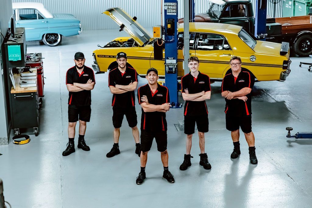 Five mechanics in black uniforms standing in a garage beside yellow and blue classic cars — Mechanic in Wollongong, NSW