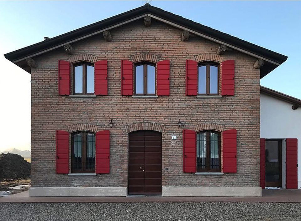 Un edificio in mattoni a due piani con persiane rosse alle finestre e un portone centrale ad arco, stagliato contro un cielo azzurro limpido.