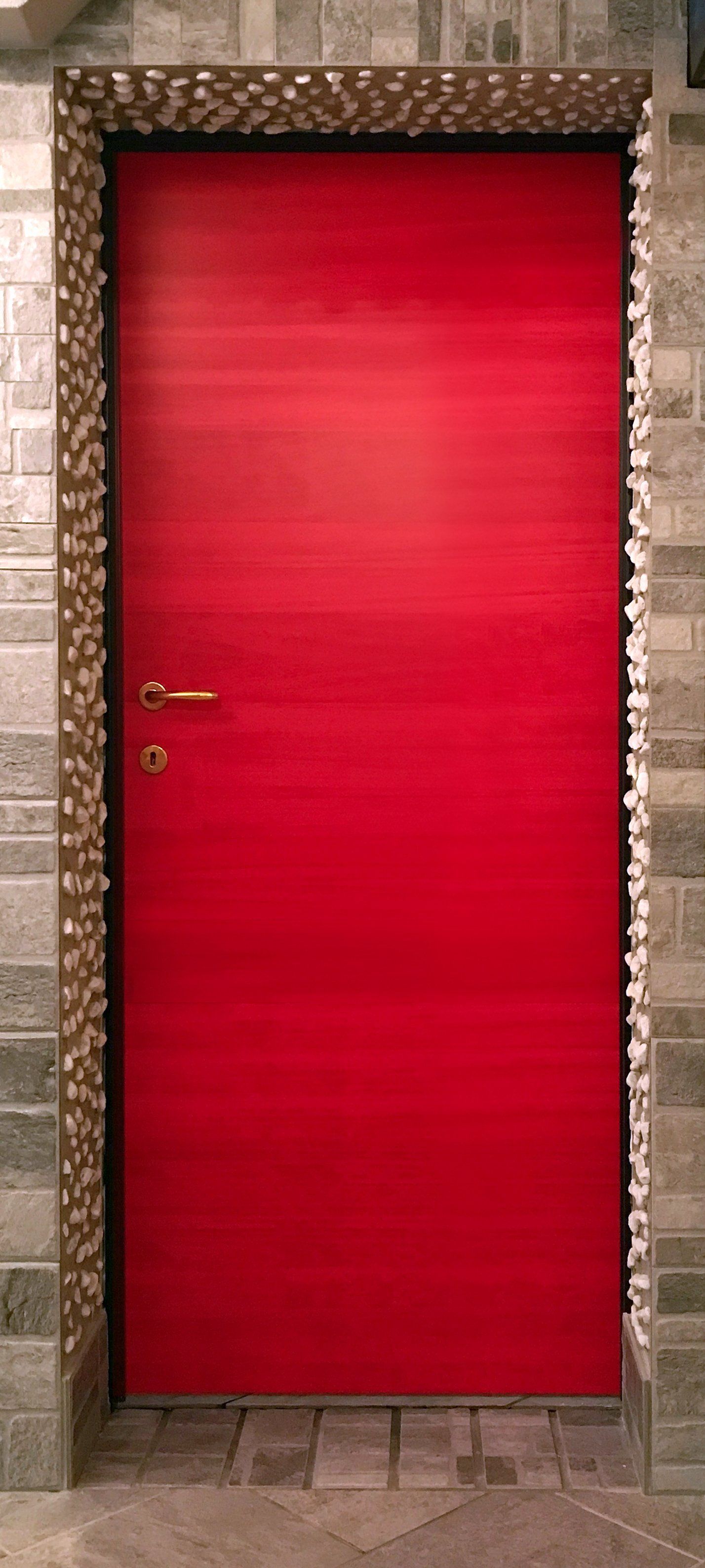 Una porta rettangolare di un rosso acceso con una semplice maniglia dorata, incorniciata da piastrelle decorative con texture su un muro di pietra.