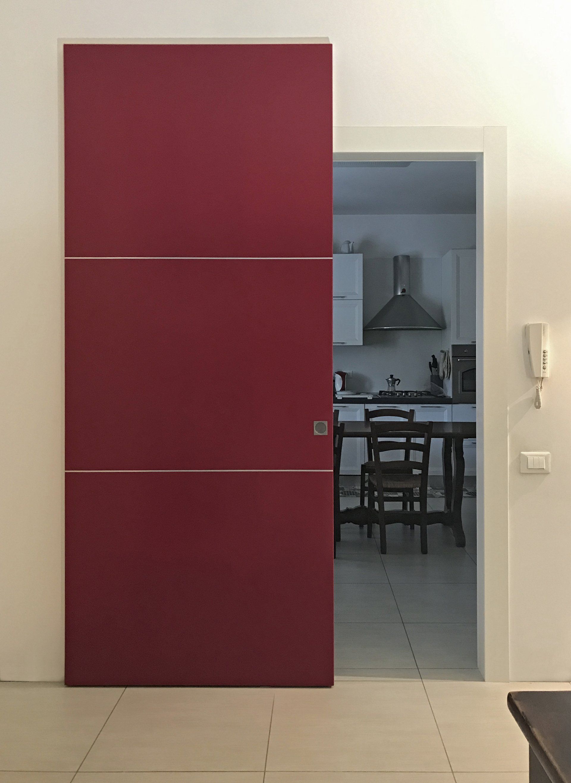 Una porta scorrevole color bordeaux opaco con due strisce orizzontali argentate, posizionata accanto a una porta che conduce alla cucina.
