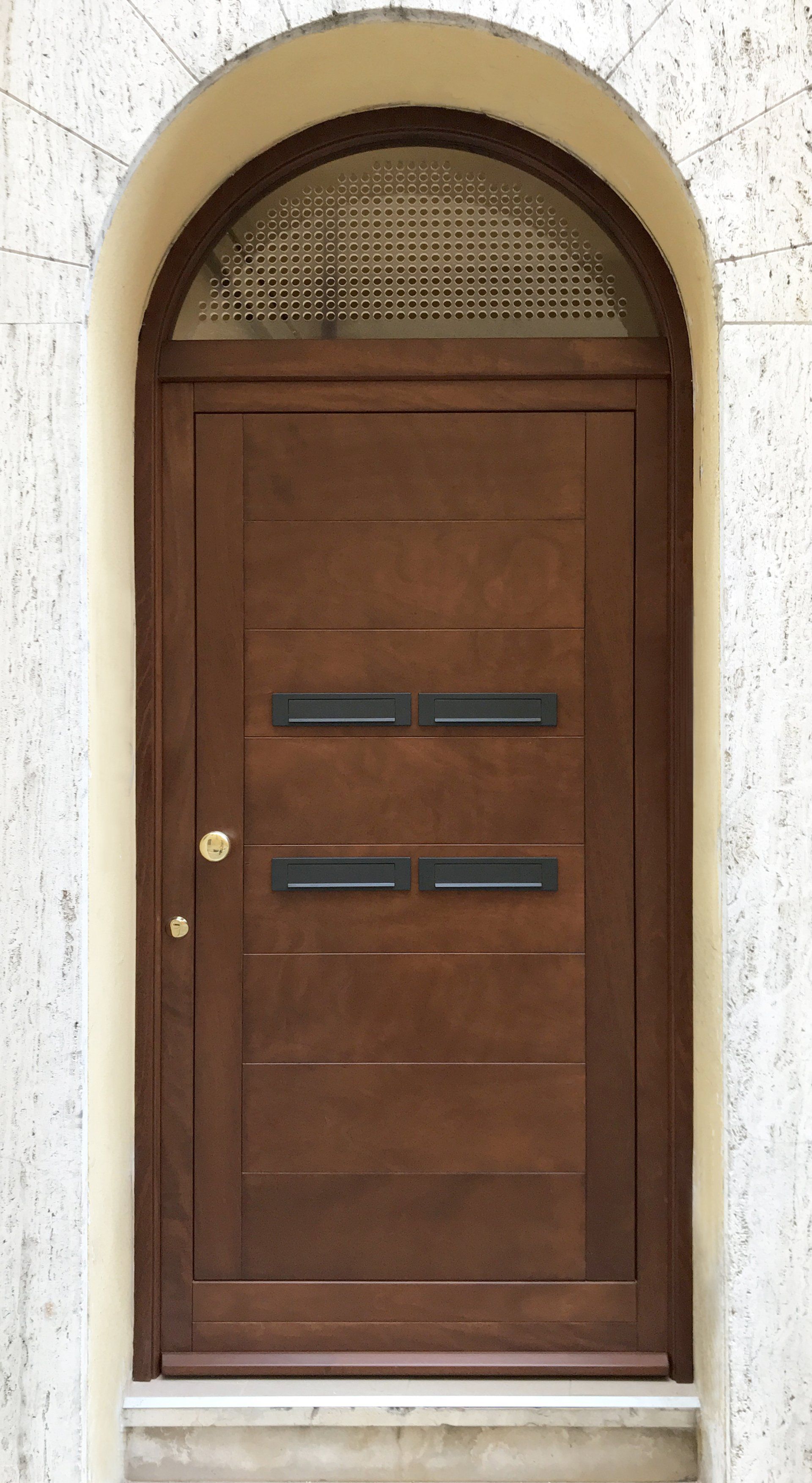 Una porta ad arco in legno scuro con due serie di fessure orizzontali per la posta, incastonate in una cornice di pietra chiara e dalla texture particolare.
