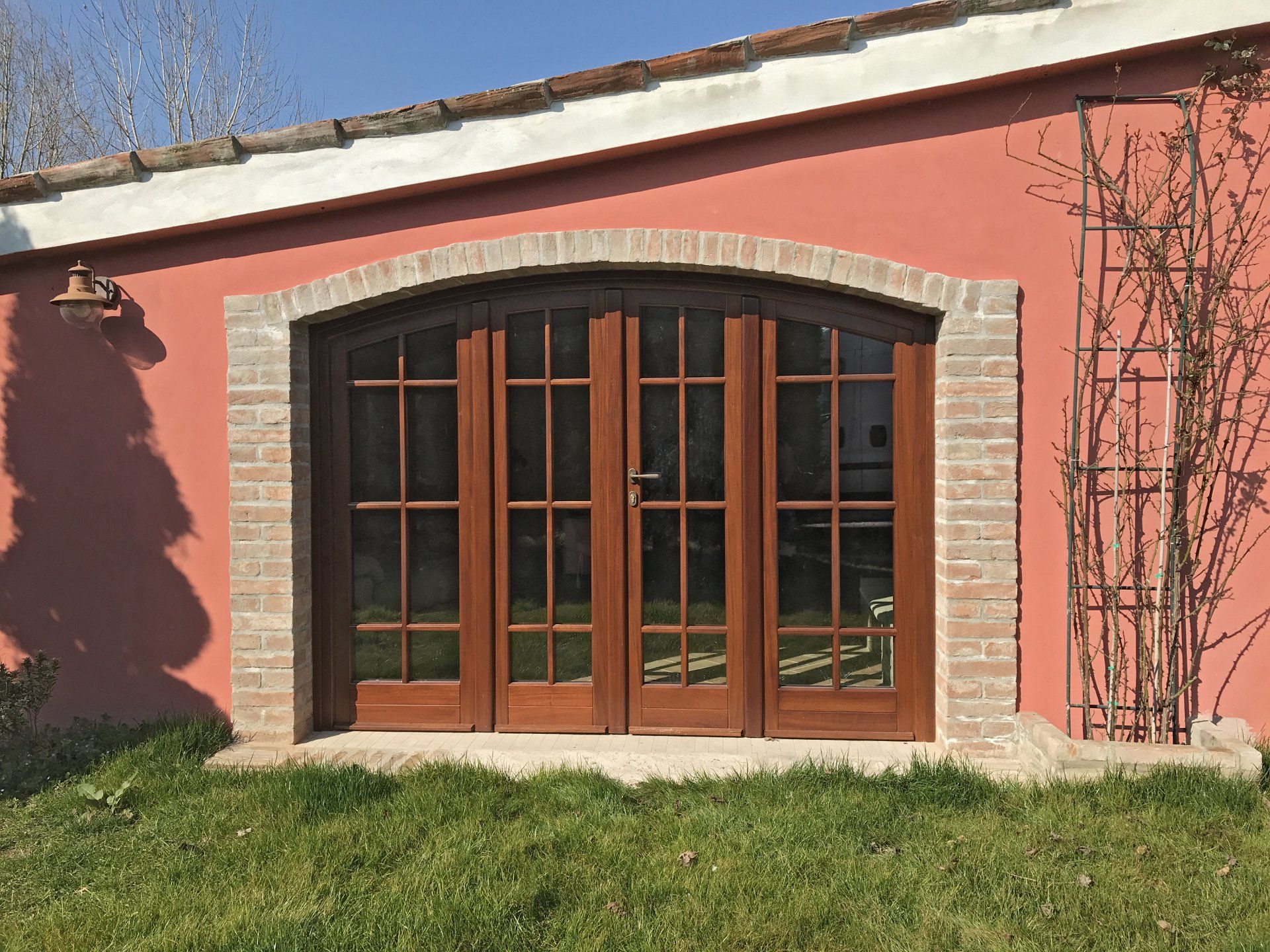 Doppie porte finestre in legno con pannelli di vetro inserite in un arco di mattoni su una parete intonacata di rosa, con un traliccio metallico sulla destra.