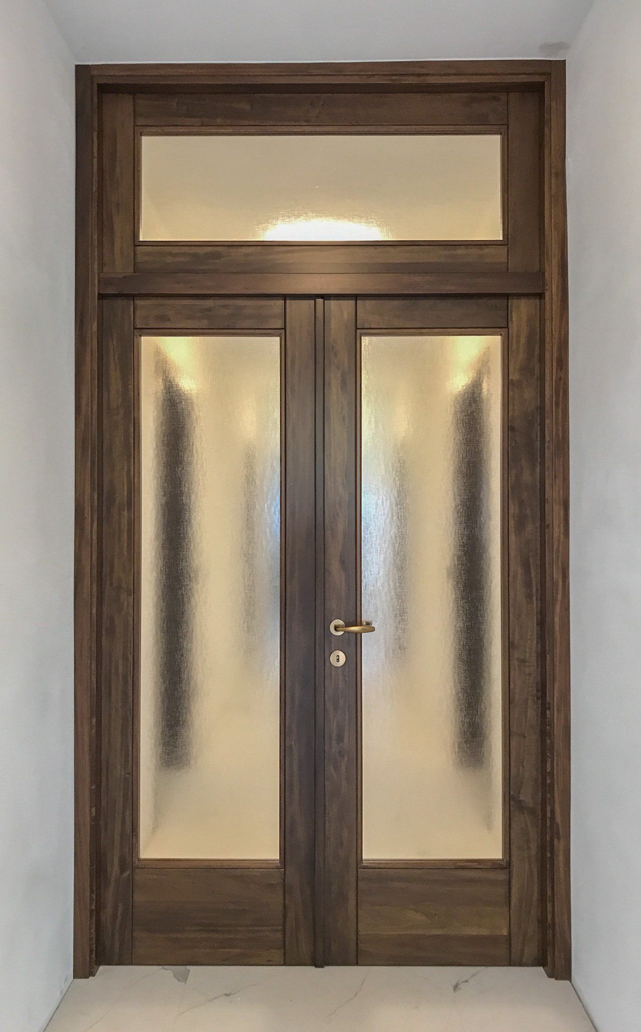 Una coppia di doppie porte interne con telaio in legno scuro venato, pannelli in vetro testurizzato e sopraluce rettangolare coordinato.