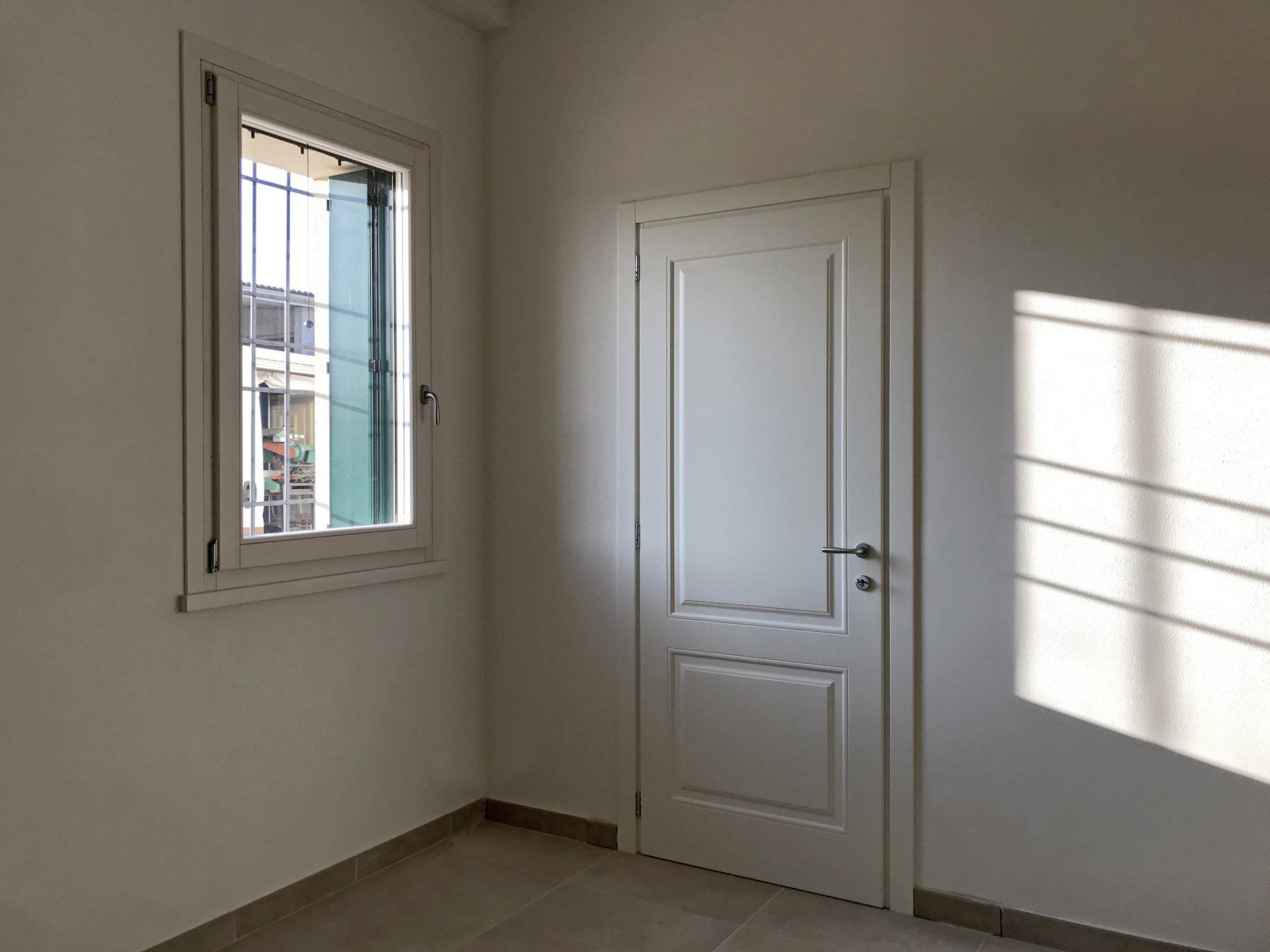 Una porta bianca e una finestra su una parete spoglia, illuminate dalla luce naturale che proietta ombre nella stanza.