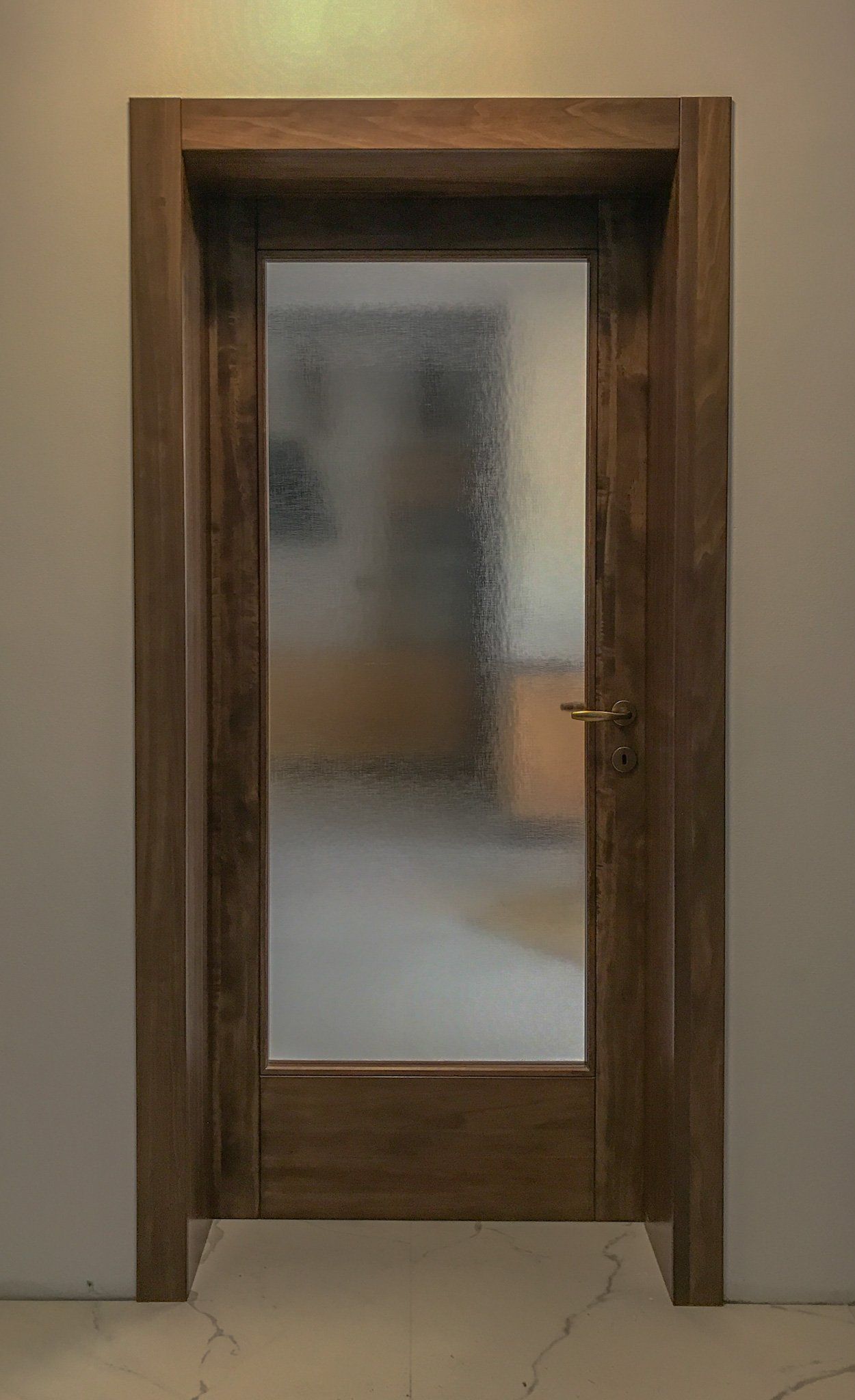Una porta in legno marrone con un pannello centrale in vetro satinato e una maniglia metallica, incastonata in una parete liscia.