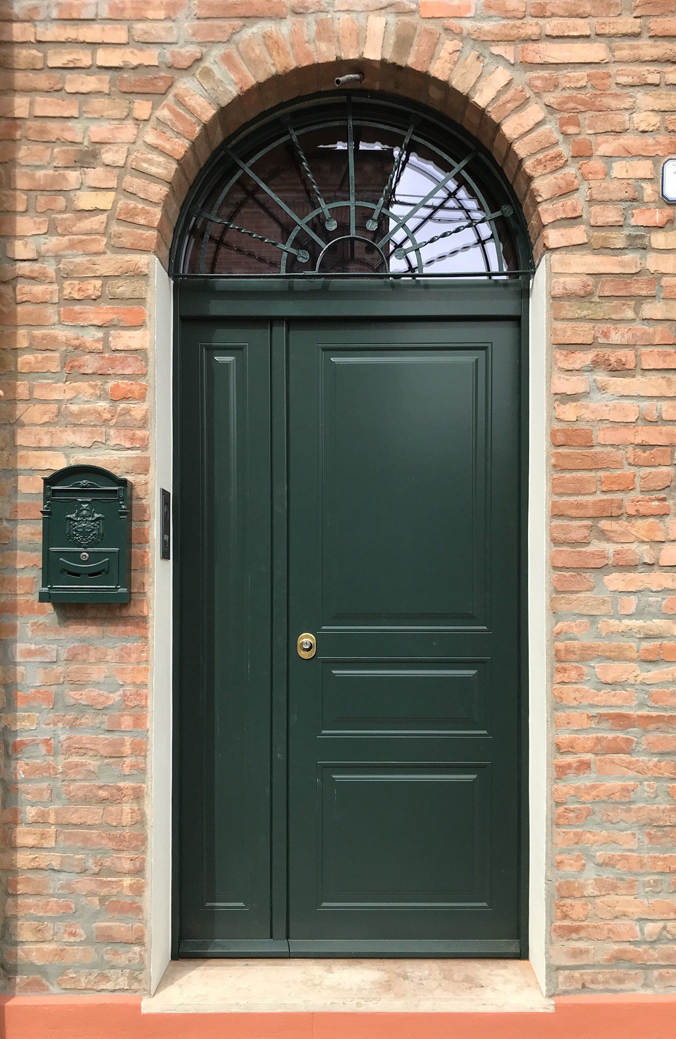 Una porta in legno verde scuro con un pannello laterale e una finestra ad arco con grata metallica, incastonata in un muro di mattoni rustici con una cassetta delle lettere abbinata.