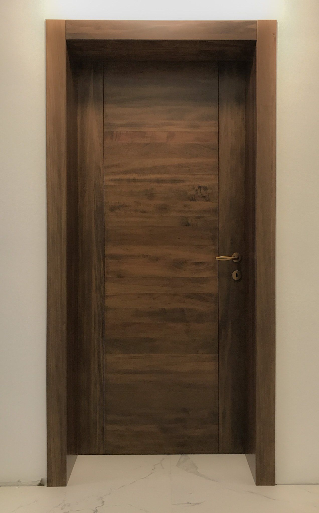 Una porta di legno chiusa, con venature orizzontali scure e una semplice maniglia dorata, incorniciata da pareti chiare.