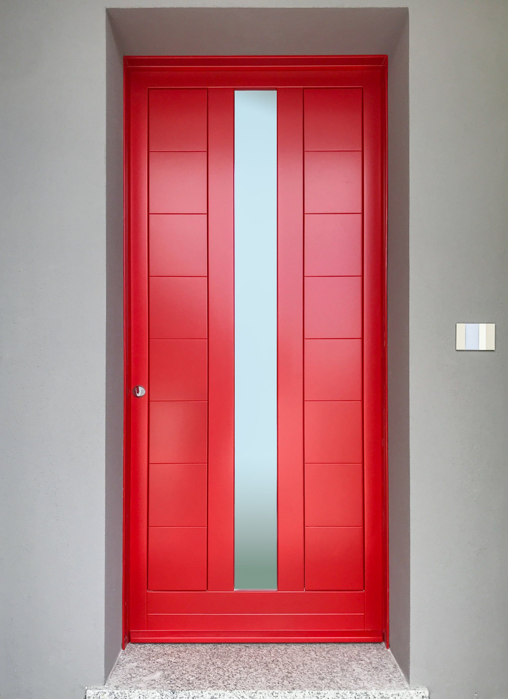 Una porta d'ingresso di un rosso acceso con un pannello verticale in vetro satinato su una parete grigio chiaro.