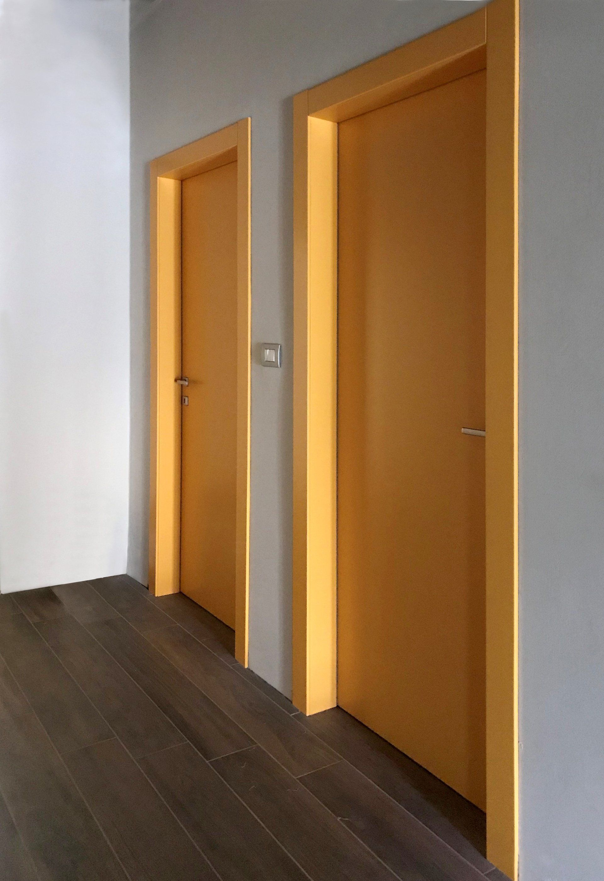 In un corridoio minimalista, due porte in legno chiaro incastonate in una parete grigia sopra un pavimento in legno scuro.