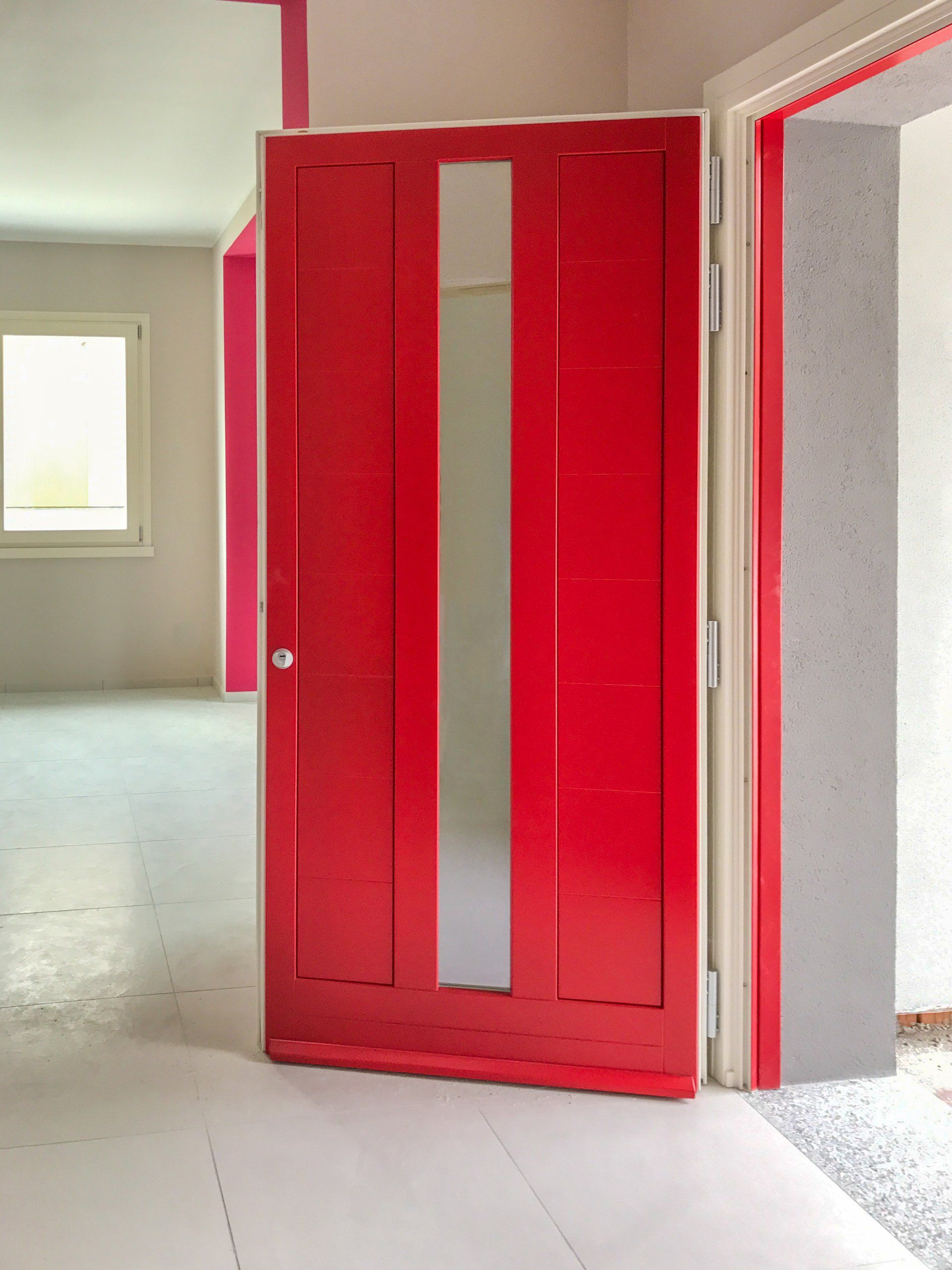 Una porta rosso acceso con un pannello centrale verticale in vetro, installata in una stanza con pavimento e pareti chiari.