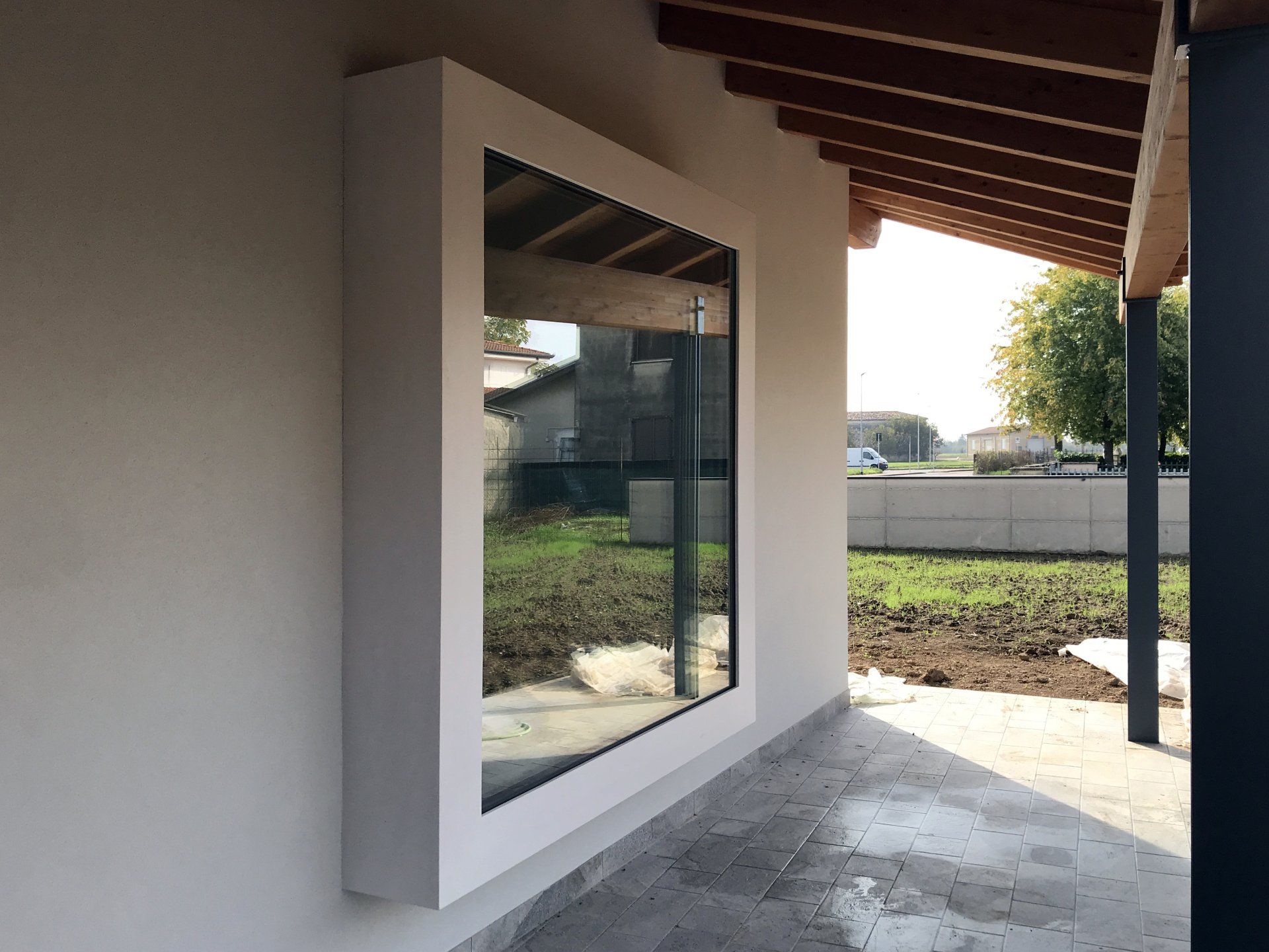 Una finestra bianca con cornice quadrata sporge da una parete esterna beige, sotto la tettoia in legno di un portico.