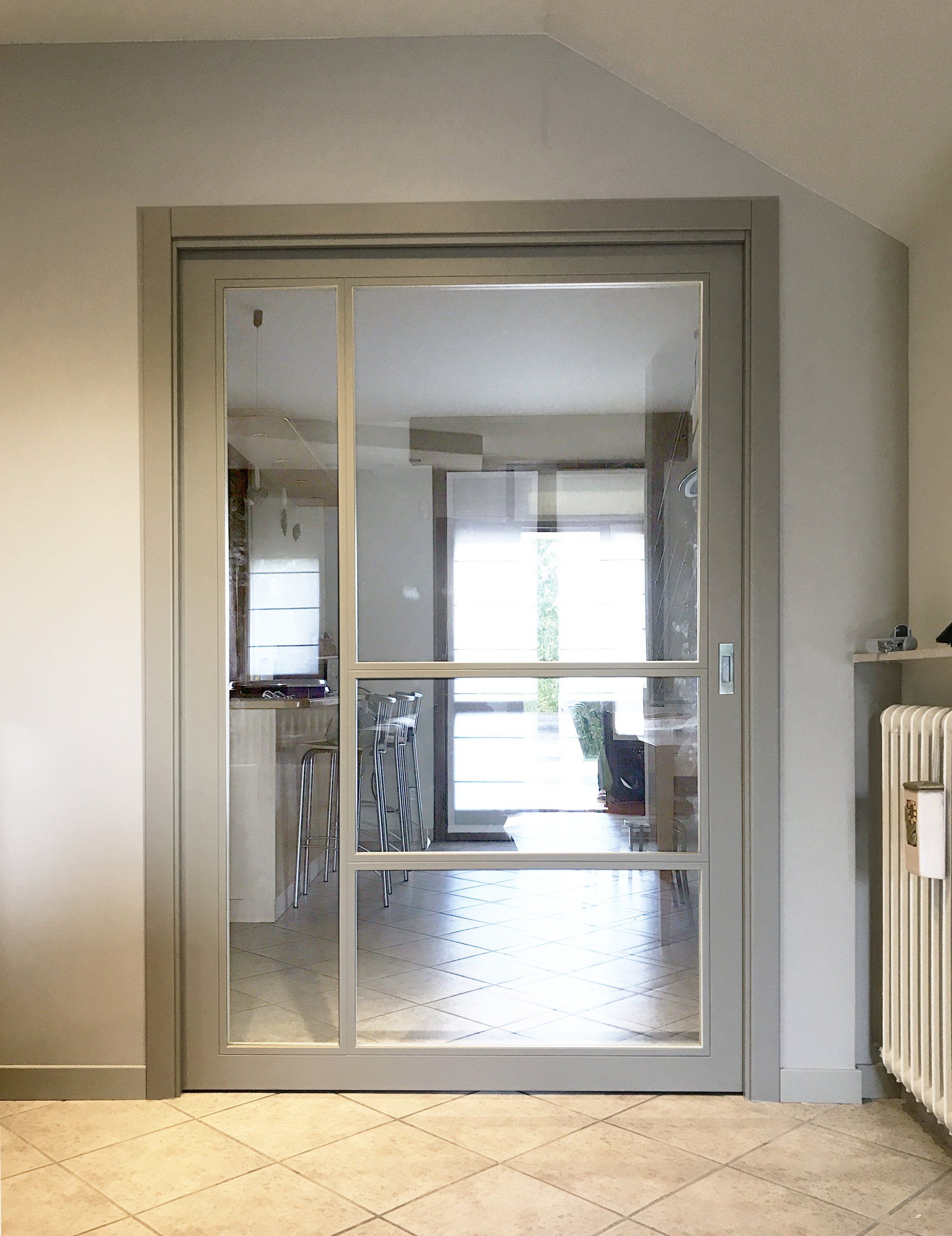 Una porta interna in vetro con cornice grigia e tre pannelli orizzontali, vista da una stanza con pavimento piastrellato chiaro che si affaccia sulla cucina.