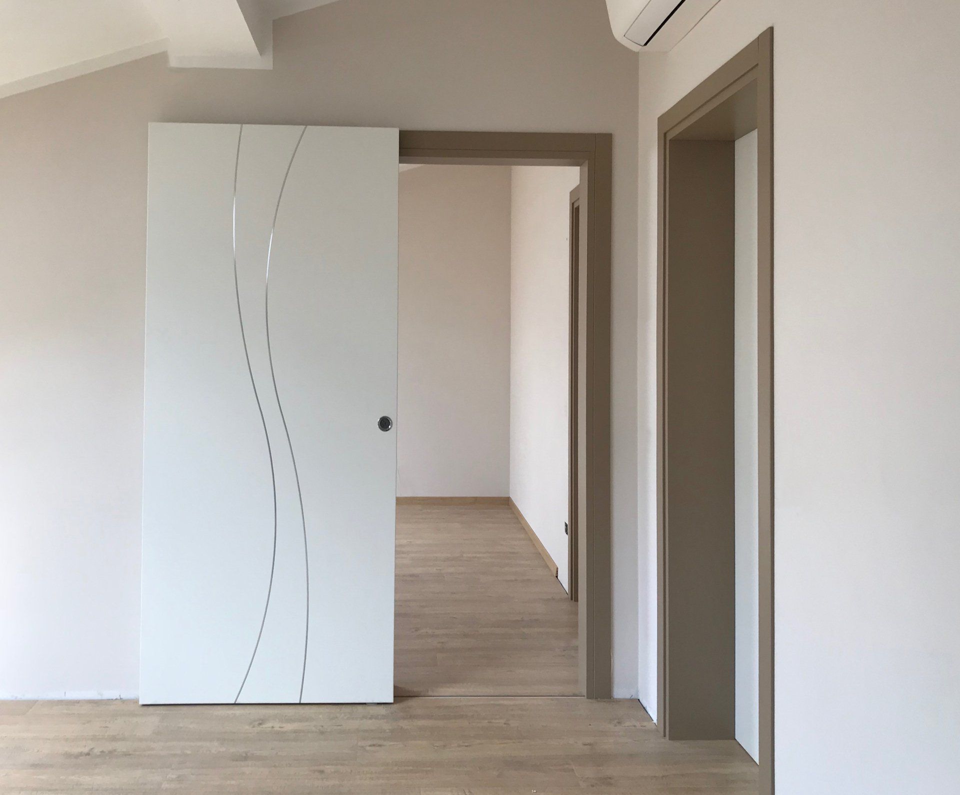 Porta scorrevole bianca con un motivo ondulato argentato, accanto a un telaio beige, in una stanza con pavimento chiaro.
