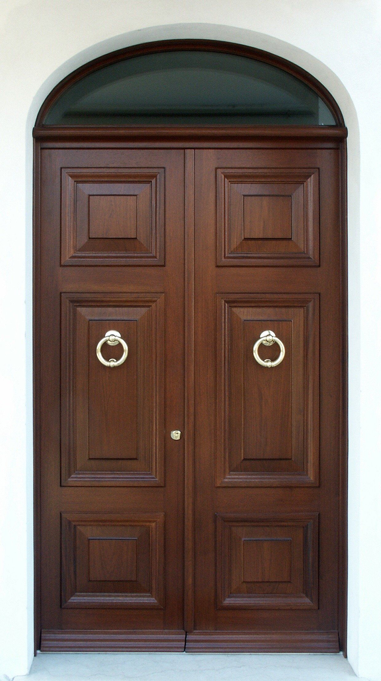 Una coppia di doppie porte in legno scuro con battenti ad anello argentati, inserite al centro in una cornice bianca ad arco con sopraluce in vetro.