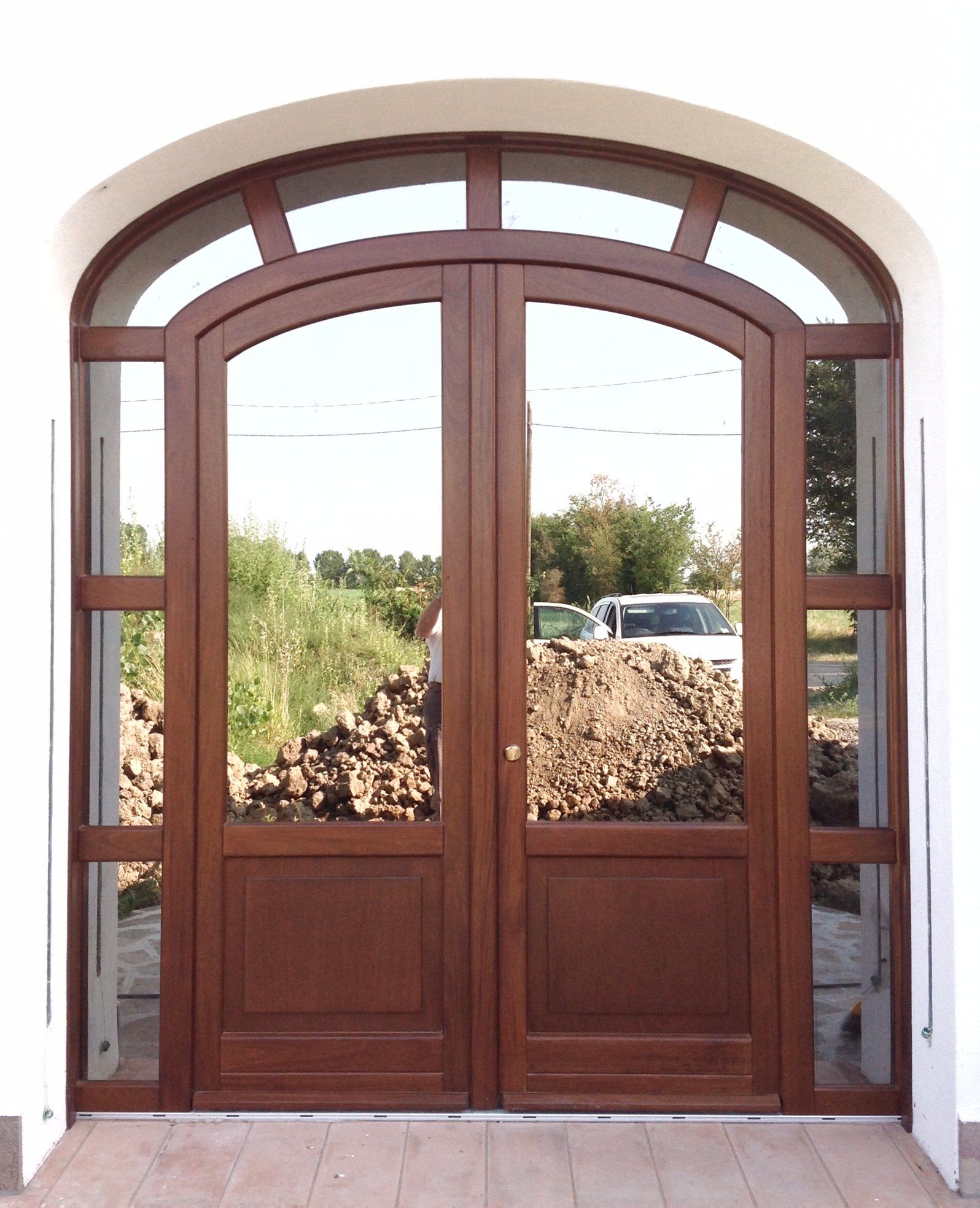 Doppia porta d'ingresso ad arco in legno con pannelli in vetro e finestre laterali, incastonata in una parete esterna bianca.