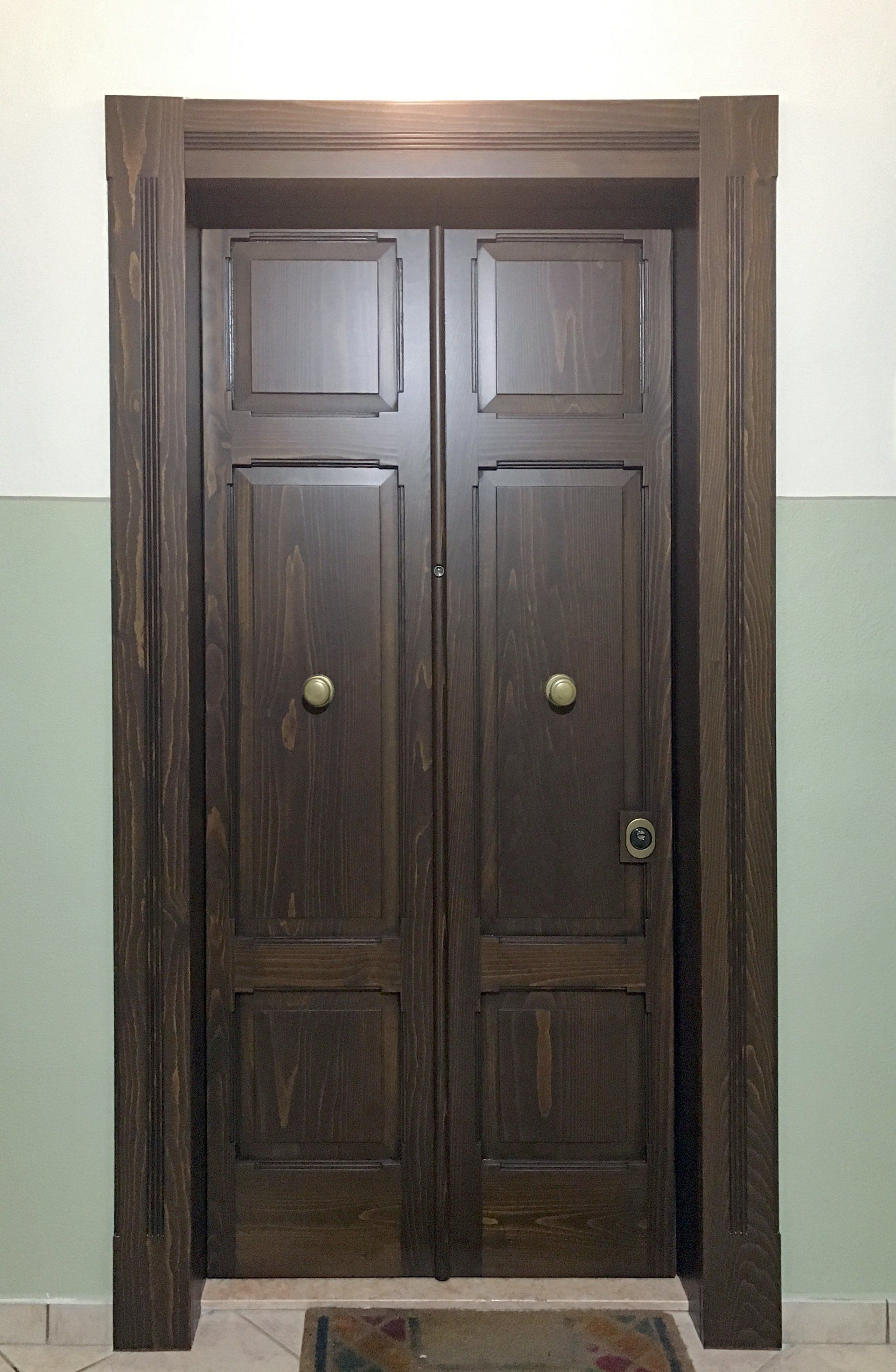 Una coppia di doppie porte in legno scuro con pomelli in ottone, inserite in un telaio su una parete dipinta di verde chiaro e bianco.