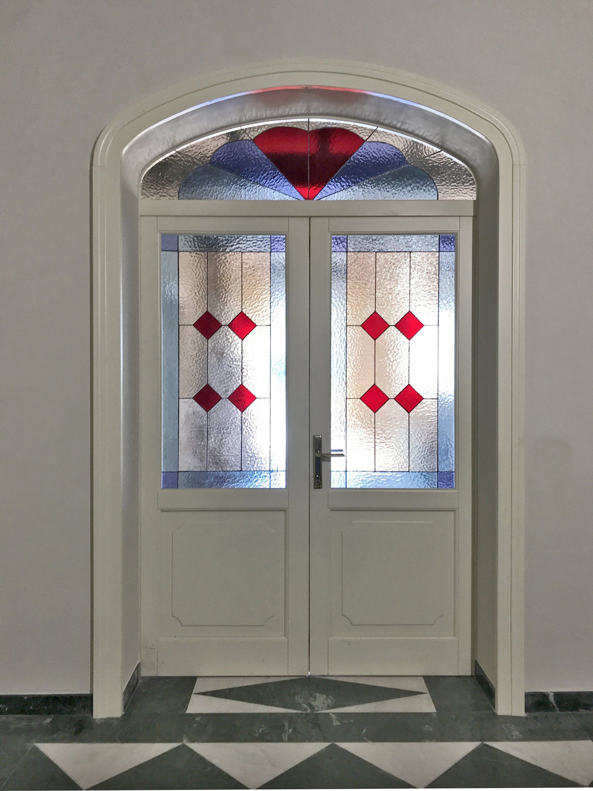 Una porta a due ante bianca con inserti a forma di diamante in vetro rosso e una finestra a lunetta semicircolare abbinata con un disegno a cuore rosso.