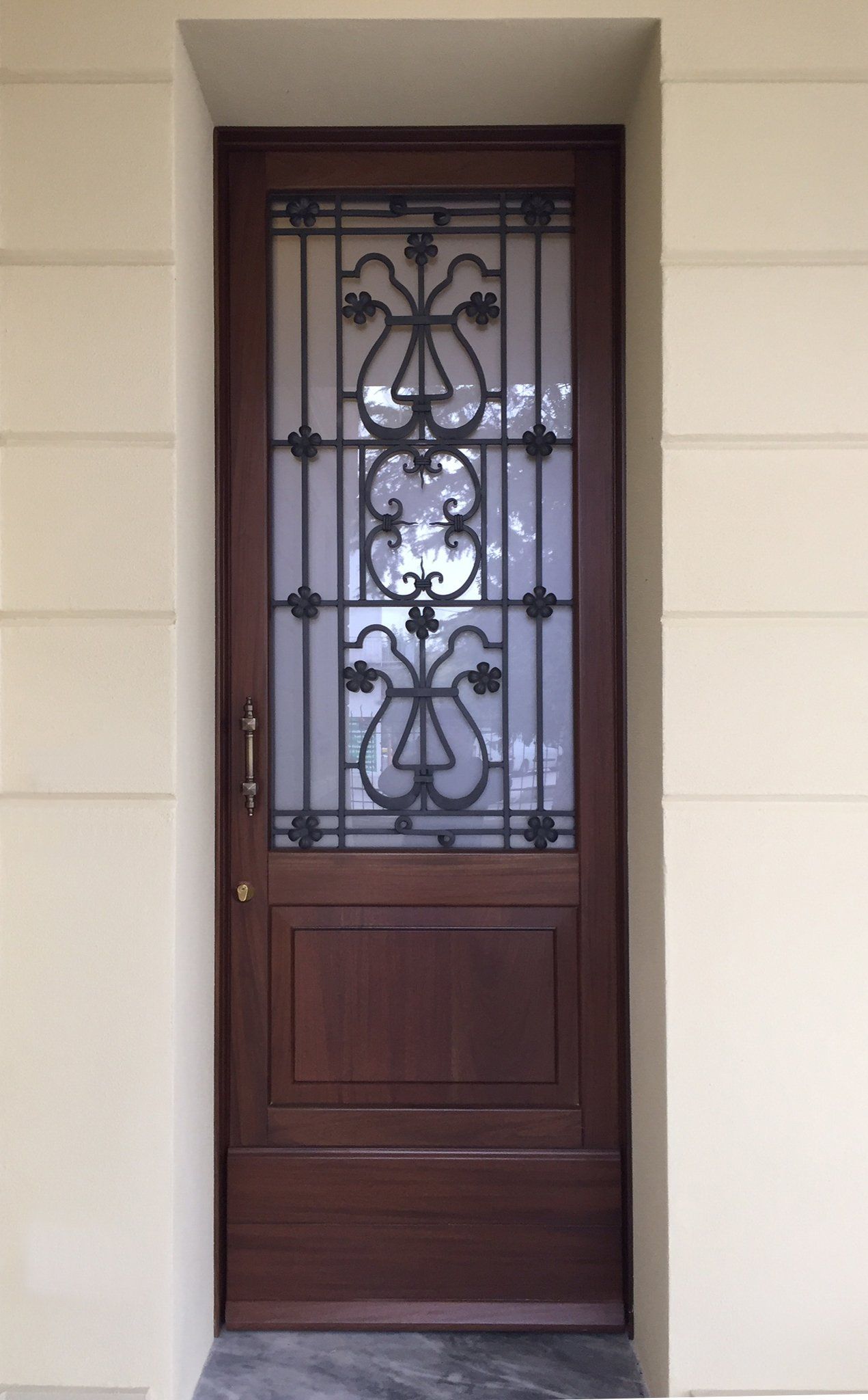 Una porta in legno marrone con una finestra centrale in vetro smerigliato decorata con elaborate volute in metallo nero e un pannello di base pieno.