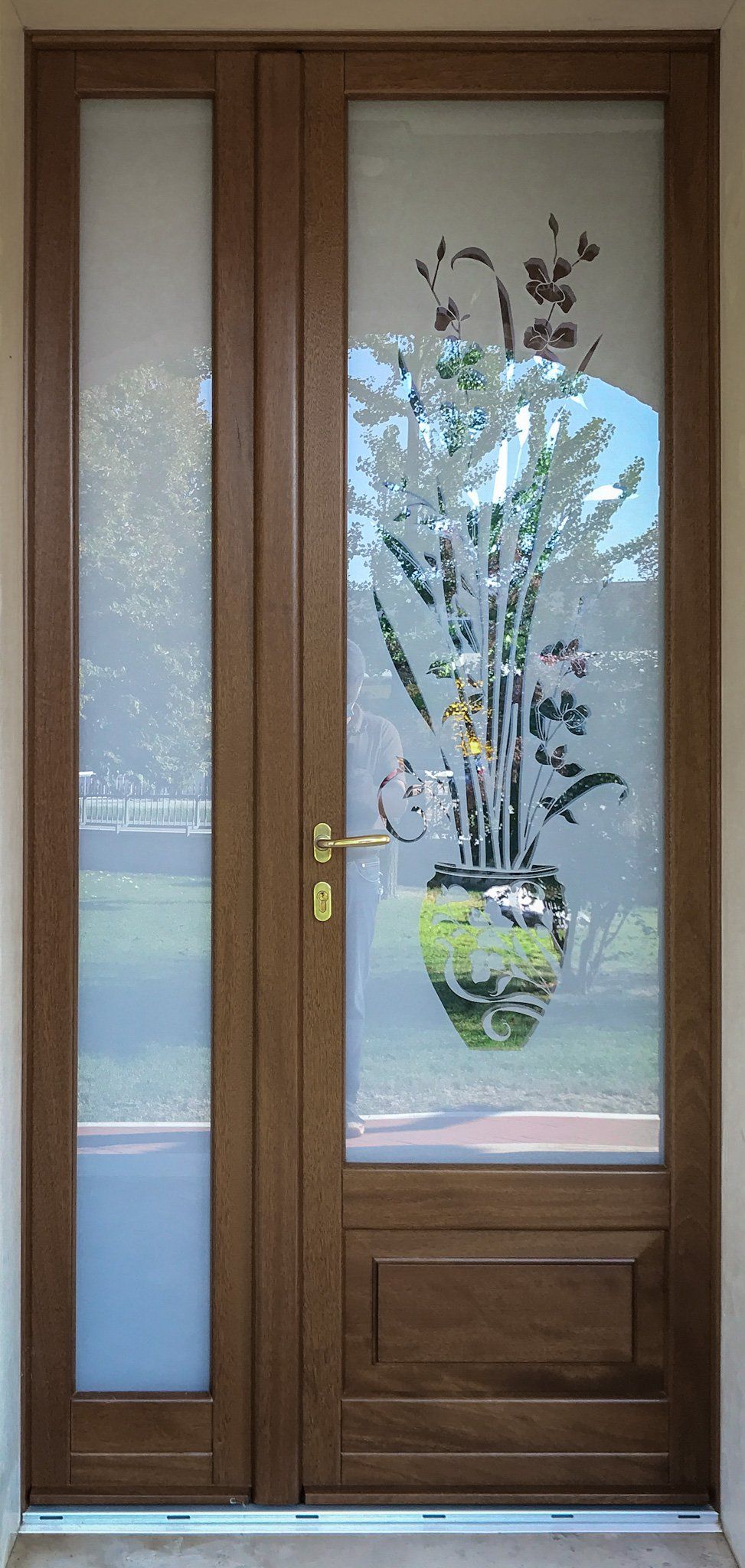 Una porta in legno con una finestra laterale rettangolare, caratterizzata da un pannello di vetro inciso con un motivo floreale e una maniglia dorata.