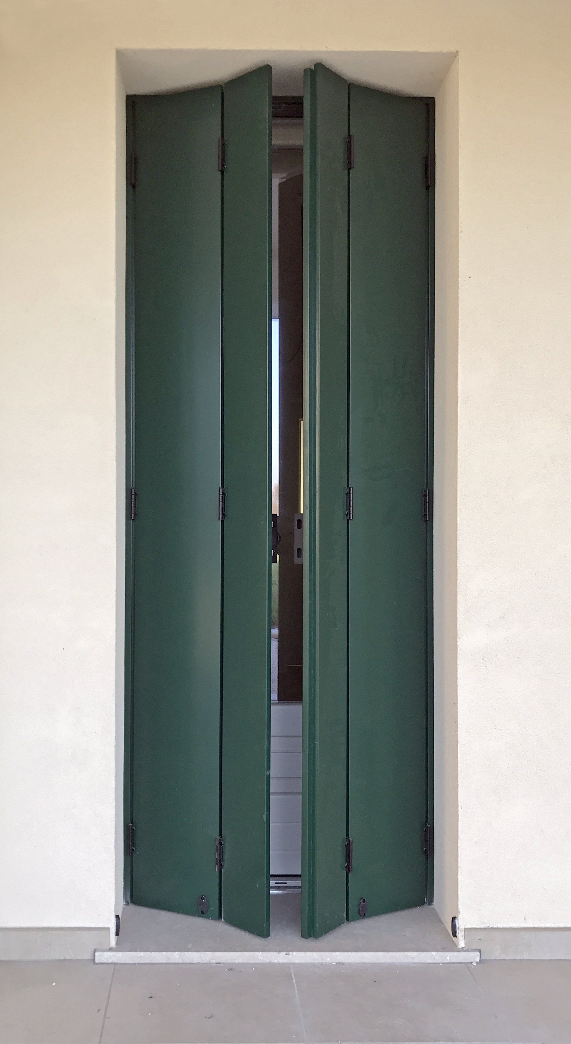 Una coppia di persiane a libro verde scuro, parzialmente aperte, montate su una cornice a muro color bianco sporco.